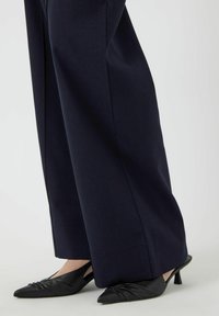 Pantalons larges bleu marine avec une texture lisse et un design épuré, associés à des escarpins à talon aiguille noirs à bride arrière avec des détails froncés.