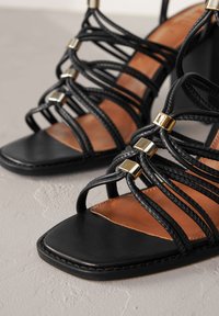 Sorte høje sandaler med stropper, der har en firkantet tådesign, guld accenter og en kombination af læder- og vævede stropper.