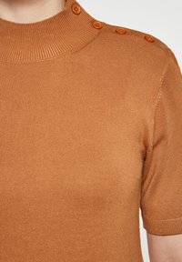 Lyhythihainen toppi lämpimän ruskeassa sävyssä, jossa on ribbireunus ja neljä oranssia nappia yksityiskohdaksi sekä sileä, teksturoitu pinta.