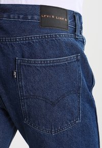 Primer plano del bolsillo trasero de unos jeans de mezclilla azul con la etiqueta negra de Levi's y "LEVI'S LINE 8" en el parche de cuero encima de la cinturilla.