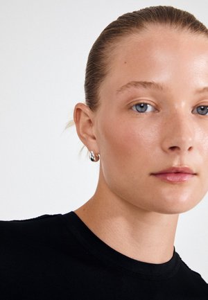 Czarny t-shirt z krótkim rękawem o gładkiej fakturze. Zbliżenie na twarz z jasną skórą, niebieskimi oczami i srebrnym kolczykiem w kształcie koła.
