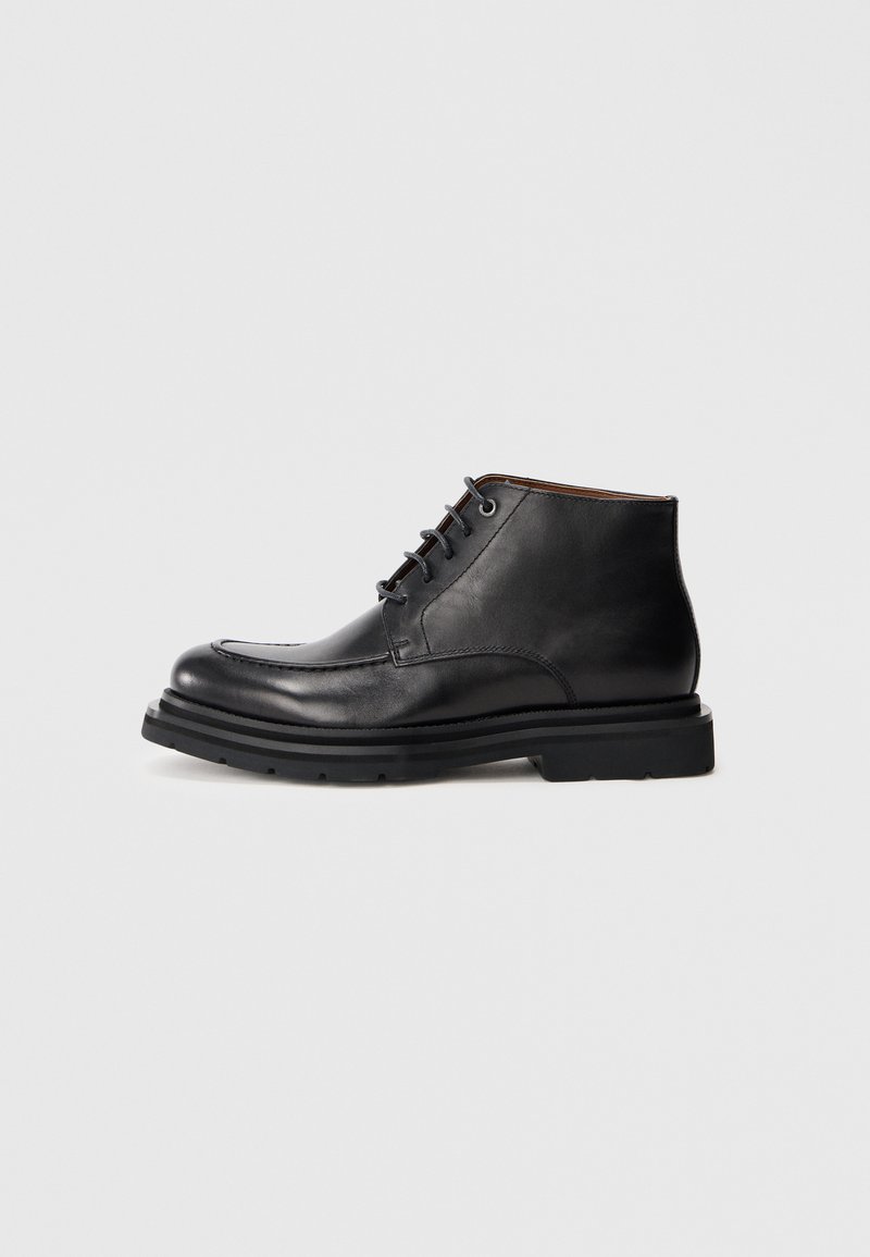 Botte en cuir noir à la cheville avec fermeture à lacets, bout rond et semelle en caoutchouc épaisse sur un fond blanc uni.