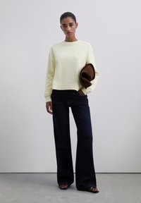 Mujer de pie contra una pared blanca, vistiendo un sudadera color crema, jeans oscuros de pierna ancha, sandalias negras, sosteniendo una chaqueta marrón doblada bajo el brazo.