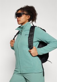 Veste en polaire teal avec une fermeture éclair à l'avant, un col montant et deux poches latérales. Portée avec un sac à dos noir et des lunettes de soleil de protection.