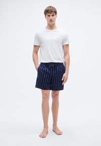 Λευκό μπλουζάκι με στρογγυλή λαιμόκοψη, συνδυασμένο με ναυτικά σορτς κολύμβησης σε σκούρο μπλε χρώμα, με κάθετες λευκές ρίγες και ελαστική μέση με κορδόνι.