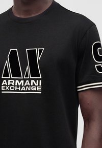 Μαύρο μπλουζάκι με έντονο λευκό λογότυπο "Armani Exchange" μπροστά και λευκή ρίγα στο μανίκι με τυπωμένο τον αριθμό εννιά.