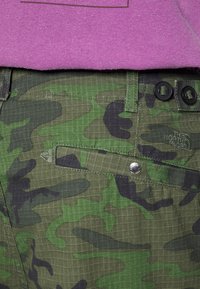 Maastokuvioidut housut, joissa vihreän ja mustan sävyjä, ruudullinen tekstuuri, kaksipainikkeinen suljenta ja kangasmerkintä kohopainettuna.