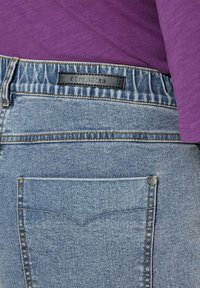 Zbliżenie na tylnią kieszeń i pas niebieskich dżinsów z czarną etykietą z napisem „#TrueSize” u góry. Włożona fioletowa koszula.