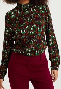 Färgglad blus med hög hals, långa ärmar och ett livligt abstrakt mönster i gröna, vinröda, bruna och vita nyanser.