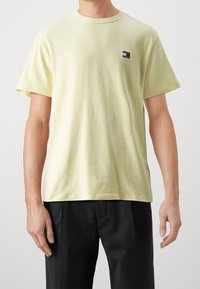 Light yellow cotton t-shirt with a round neck and short sleeves. Features a small multi-colored logo patch on the left chest.