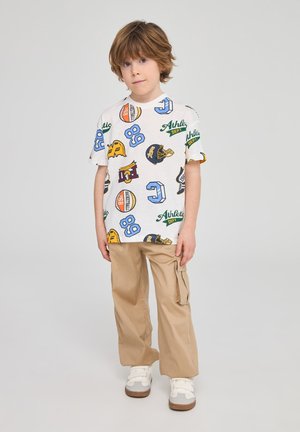 Maglietta bianca con loghi sportivi colorati e testo, abbinata a pantaloni cargo beige con tasche laterali e scarpe da ginnastica bianche con accenti grigi.