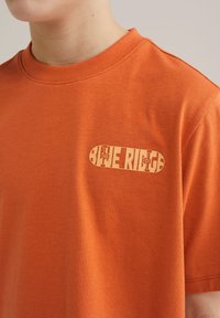 Pomarańczowy bawełniany t-shirt z logo z nadrukiem w kolorze beżowym. Okrągły dekolt, krótkie rękawy i luźny krój. Gładka tekstura.
