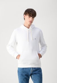 Weißer Baumwollhoodie mit Kängurutasche, Kapuze mit Kordelzug und kleinem Logo auf der linken Brust. Kombiniert mit blauen Jeans.