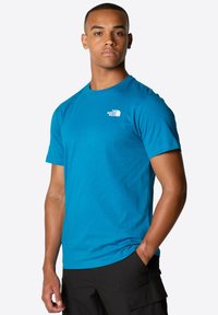 Camiseta de cuello redondo azul hecha de una tela suave, con un pequeño logo blanco en la parte superior izquierda, combinada con pantalones cortos cargo negros.