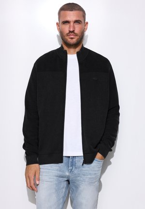 Mann mit kurzen Haaren und Bart, der eine schwarze Reißverschlussjacke, ein weißes T-Shirt und hellblaue Jeans trägt, steht vor weißem Hintergrund.