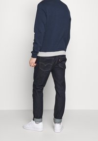 Hombre de espaldas, vistiendo un sudadera azul marino, con cinturilla gris y jeans de mezclilla oscuros con los puños doblados. Zapatillas blancas completan el atuendo.