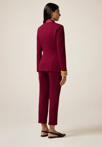 Completo bordeaux con giacca aderente e pantaloni a sigaretta. La giacca presenta un colletto in satin; abbinata a sandali neri a pianta piatta. Tessuto liscio.