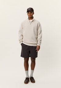 Μπεζ φλις φούτερ με φερμουάρ, σκούρες βερμούδες, γκρίζες κάλτσες, καφέ παπούτσια και σκοτεινό καπέλο με άσπρα γράμματα· απλός, casual στιλ.