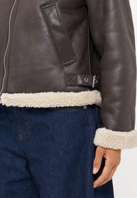 Giubbotto in pelle marrone con bordo in shearling beige. Presenta una chiusura zip, tasche laterali e un dettaglio con fibbia in vita.