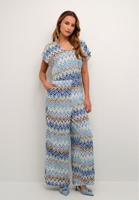 Mustriline hüpits, millel on lühikesed varrukad, V-kaelus ja taskud. Omab geomeetrilist disaini sinises, beežis ja pastelltoonides. Lahtine lõige.