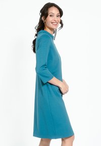 Teal klänning med trekvartslånga armar, avslappnad passform och rund hals. Tillverkad av mjukt tyg, har sidofickor och knälång nederkant.