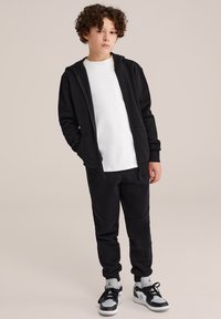 Melna zipa kapuci virs baltas T-kreklu, apvienota ar melnām sporta biksēm. Melnbalti sporta apavi. Gluda tekstūra, ikdienišķs dizains.