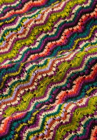 Tessuto lavorato a maglia multicolore caratterizzato da un motivo a zigzag in vivaci tonalità di verde, viola, rosa e marrone, con una lieve texture e accenti scintillanti.