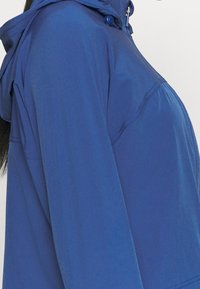 Ljusblå, vattenavvisande jacka med huva. Slät textur, dragkedjestängning och dubbla knappdetaljer vid halsen. Har sömdetaljer.