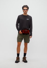 Άνδρας με γυαλιά ηλίου, μαύρο πουκάμισο με μακριά μανίκια, χακί σορτς cargo, πεζοπορικά μποτάκια και κρατώντας μία κόκκινη τσάντα μέσης μπροστά από έναν απλό φόντο.