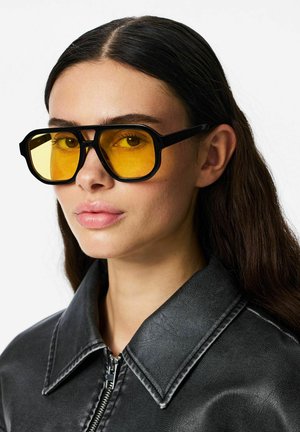 Kvinde med langt mørkt hår bærer store sort-indrammede briller med gule linser og en sort læderjakke med lynlåsåbning ved kraven.