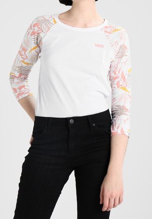Personne portant un t-shirt blanc Vans avec des manches trois-quarts à motif floral et un pantalon noir, debout avec une main sur la hanche.