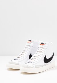 Baskets montantes blanches en cuir et daim, avec un logo swoosh noir, une semelle en caoutchouc et une marque orange sur la languette.