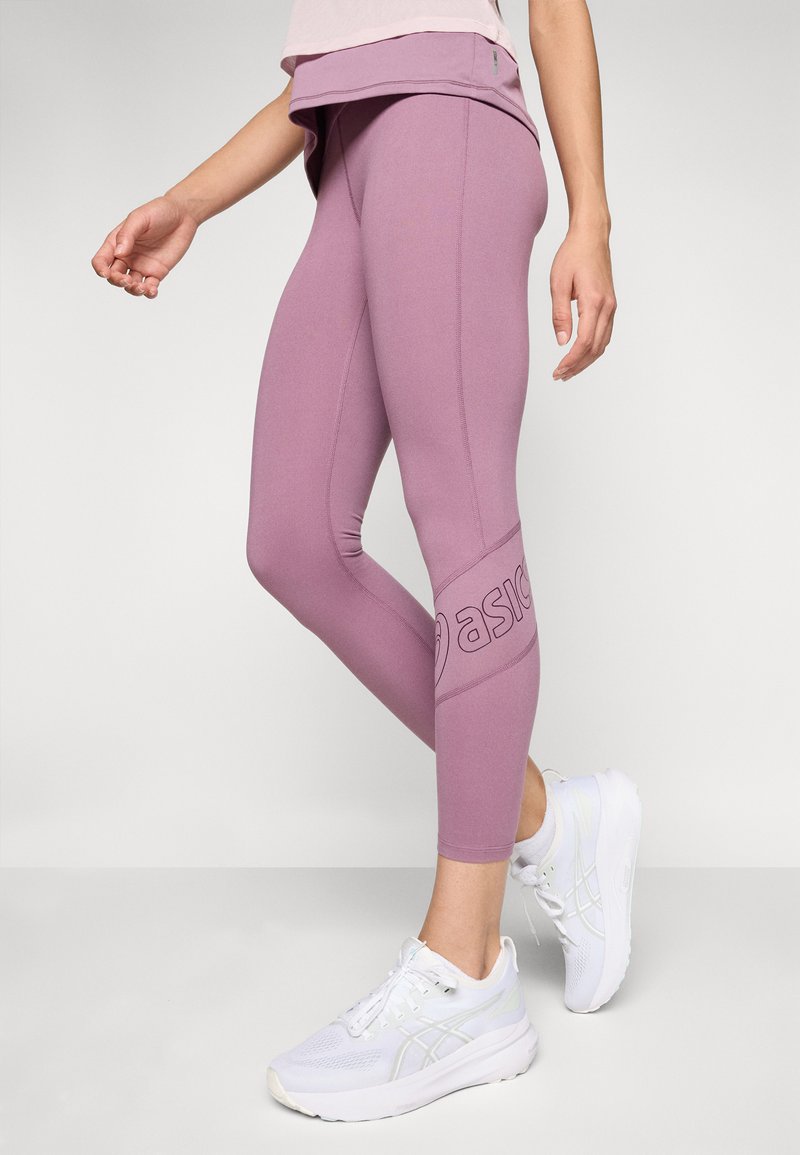 Leggings violets en tissu extensible, ornés d'un logo sur le côté, conçus pour le confort et le mouvement, associés à des chaussures de sport blanches.