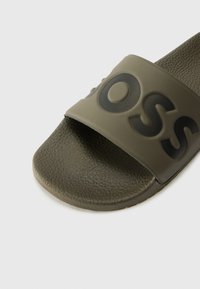 Olive green slide sandal with a textured footbed and a wide strap featuring a black embossed logo. Smooth finish and rubber sole.