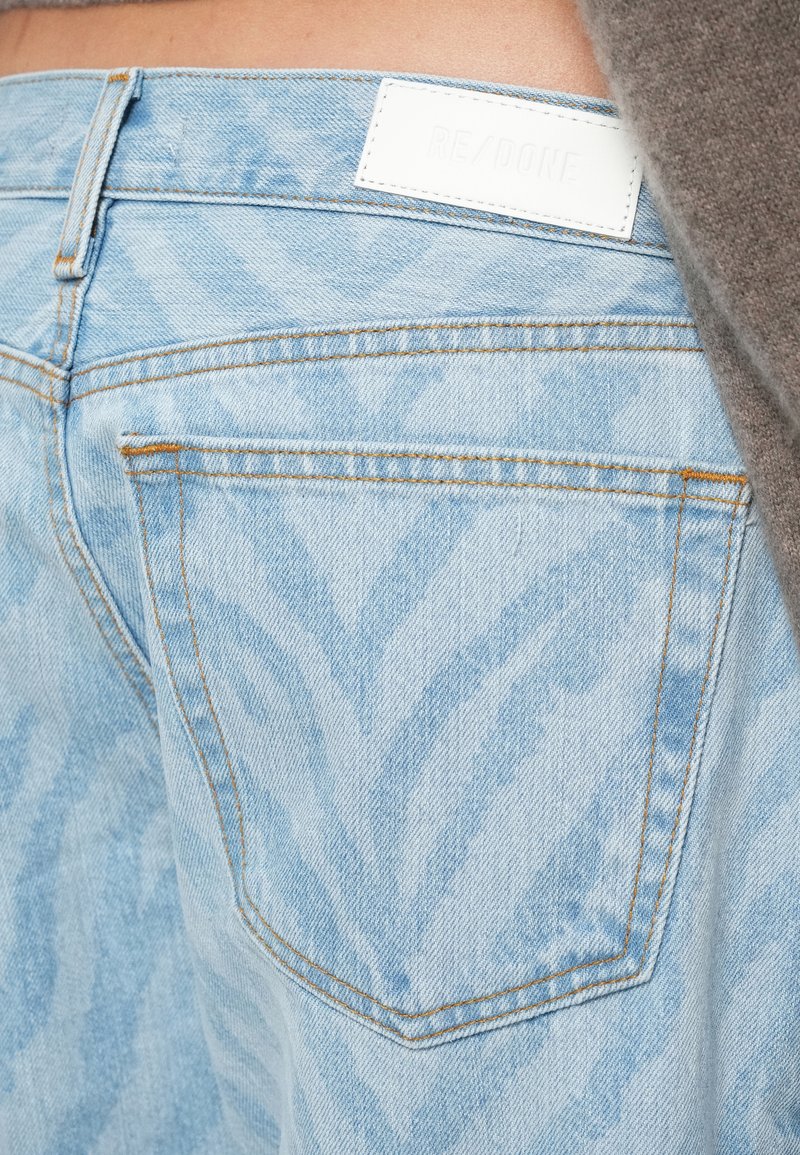 Lichtblauwe denim jeans met oranje stiksels en een wit label op de tailleband, gedragen met een bruine top.