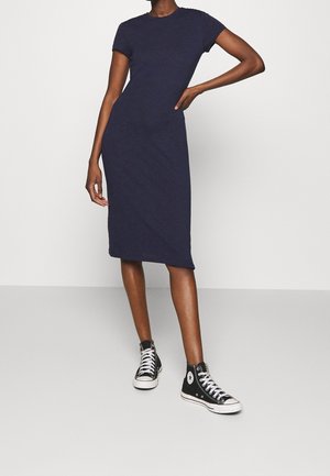 Femme portant une robe ajustée bleu marine longueur genou avec manches courtes et des baskets montantes noires, debout avec une main sur la hanche.