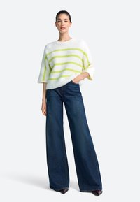 Pulover alb pufos cu dungi verzi, mâneci largi; asortat cu blugi largi, bleu închis. Modelul stă cu mâinile în buzunare.