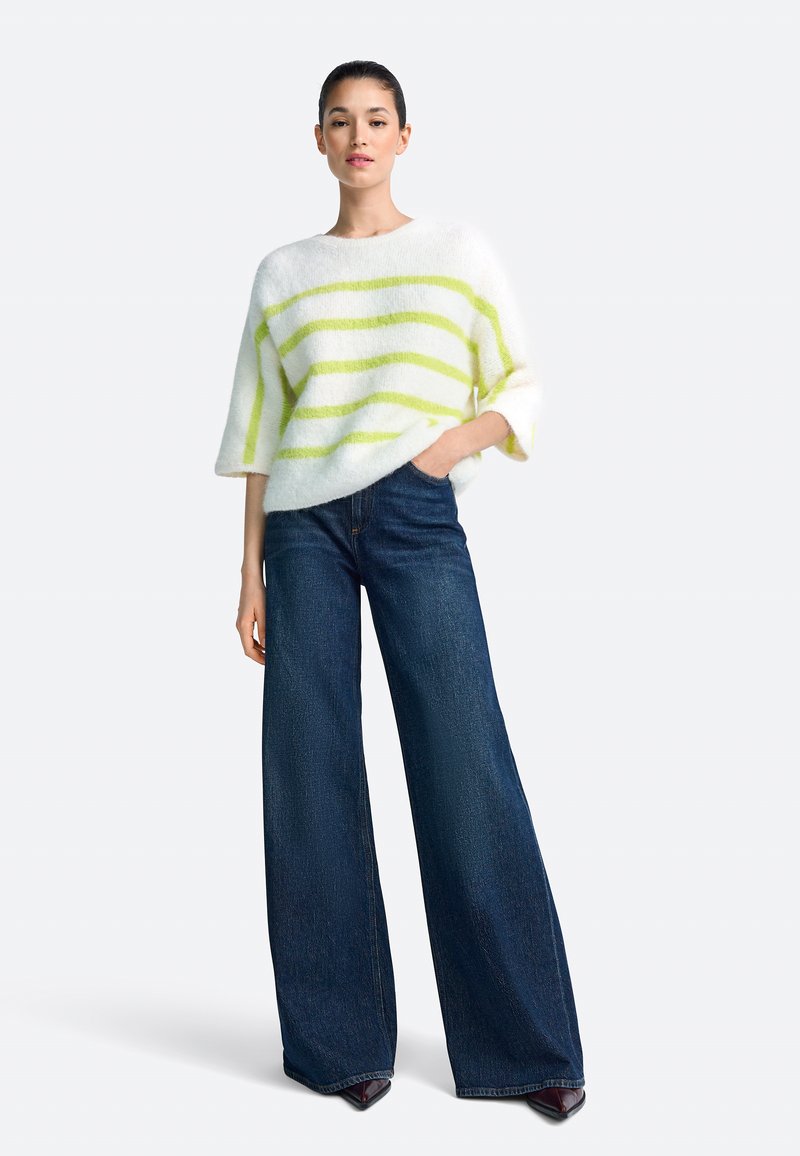 Pulover alb pufos cu dungi verzi, mâneci largi; asortat cu blugi largi, bleu închis. Modelul stă cu mâinile în buzunare.