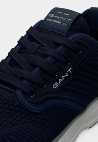 Αθλητικό παπούτσι σε ναυτικό μπλε, με υφασμάτινο διχτυωτό επάνω μέρος, επίπεδα κορδόνια και λευκή σόλα. Το λογότυπο GANT είναι εμφανώς τοποθετημένο στη γλώσσα του παπουτσιού.