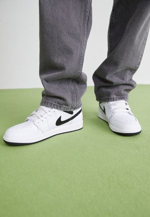 Person trägt weiße Nike-Sneaker mit schwarzem Swoosh und grauen Jeans und steht auf grünem Boden.
