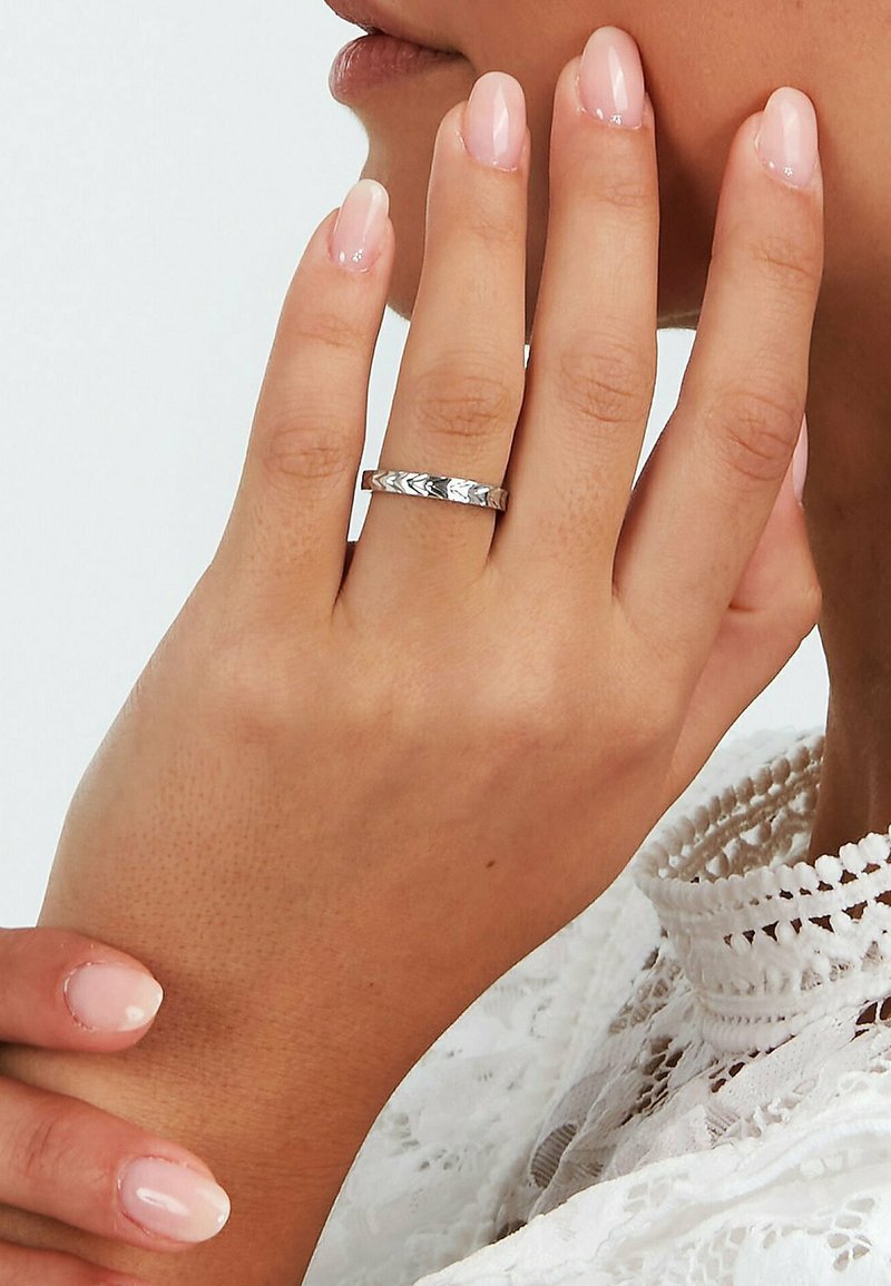 Mano con unghie rosa pallido lucide che indossa un anello argentato decorato sull'anulare, toccando il viso vicino alle labbra, indossando un indumento di pizzo bianco.