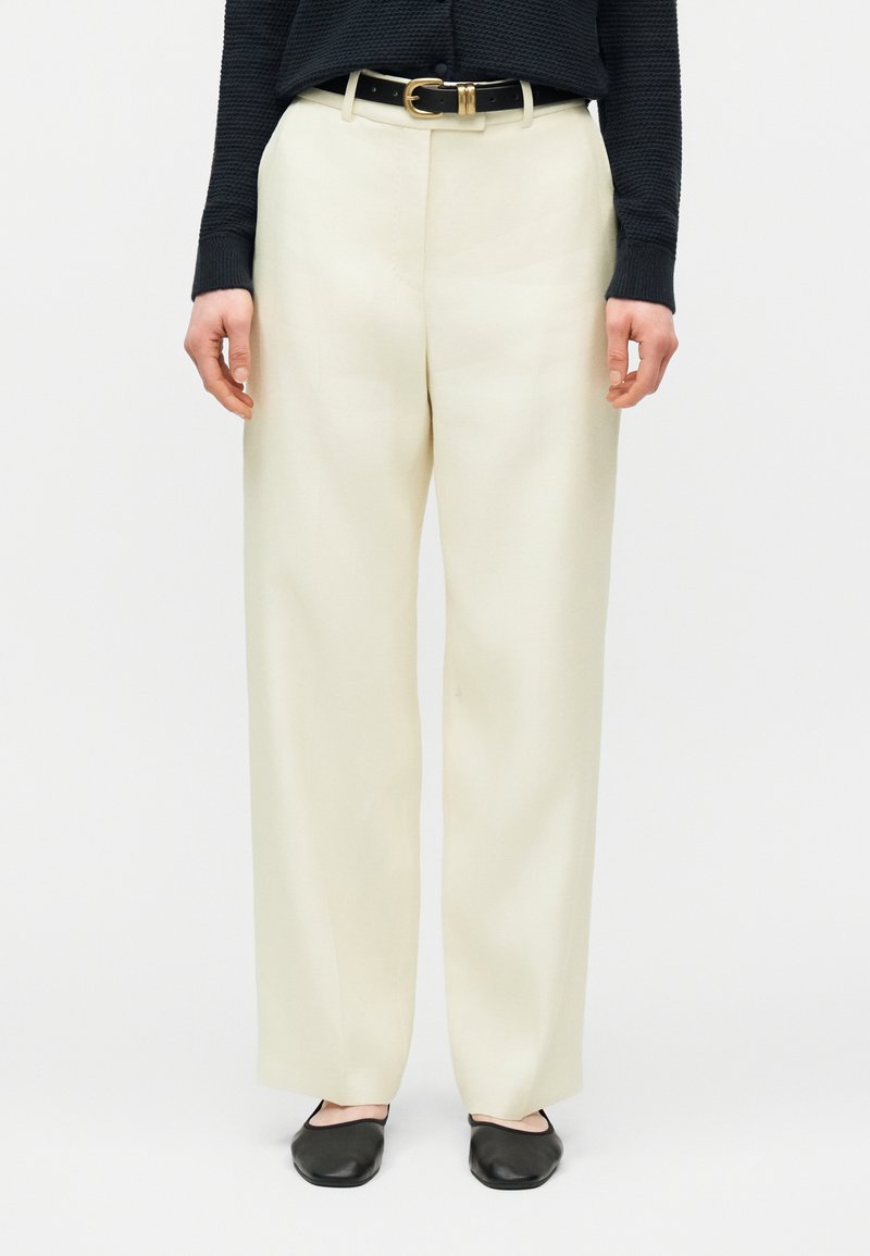 Άτομο που φοράει κρεμ παντελόνια με φαρδιά μπατζάκια, μαύρη ζώνη με χρυσή πόρπη, μαύρο μακρυμάνικο τοπ και μαύρα φλατ παπούτσια, στέκεται μπροστά σε λευκό φόντο.