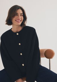 Črn kardigan z zlatimi gumbi, okroglo izrezan, sproščenega kroja in teksturirane tkanine. Model sedi na stolu z rjavim usnjenim naslonjalom.