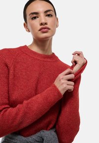 Pull en tricot rouge avec une texture côtelée, un col rond et des manches longues. Le mannequin ajuste le poignet avec une ceinture grise en dessous.