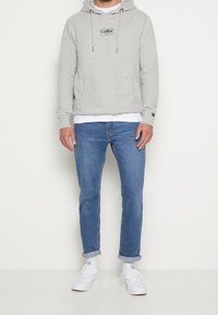 Homme portant un sweat à capuche gris clair, un t-shirt blanc, un jean bleu retroussé, des chaussettes blanches et des baskets blanches, debout devant un fond blanc.