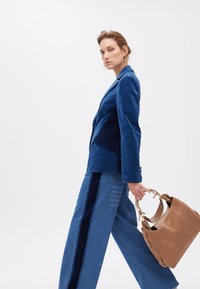 Una modella cammina con sicurezza, indossando un blazer di velluto a coste blu e jeans a gamba larga, tenendo una borsa color cammello, su uno sfondo bianco.