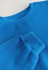 Blauer Pullover aus gestricktem Stoff mit rundem Ausschnitt und gerippten Bündchen. Glatte Textur und gleichmäßige Farbung im gesamten Kleidungsstück.