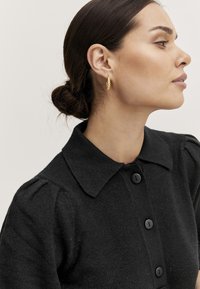Indumento in maglia nera con scollatura a colletto e chiusura con bottoni. Presenta maniche a sbuffo e una finitura testurizzata. Orecchino ad anello dorato come accento.