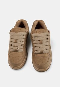Sneaker beige con tomaia in pelle liscia, punta tondeggiante e dettagli perforati. Presentano lacci piatti beige e supporto cuscinetto per la caviglia.