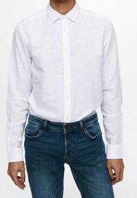Camisa blanca de manga larga hecha de lino, con cuello abotonado y tapeta frontal, combinada con unos vaqueros de mezclilla azul oscuro.